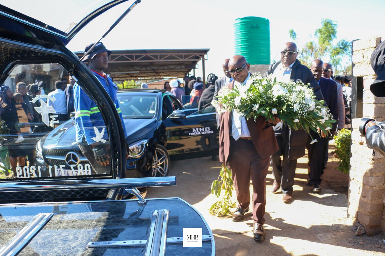 Mourners carrying a floral wreath to the MHB glass hearse trailer